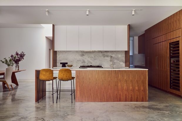 Contemporary open-plan kitchen with a large timber-clad island, white upper cabinetry, a mottled stone backsplash and benchtop, and polished concrete flooring. Two upholstered bar stools sit at the island, while a timber-lined wall with integrated storage extends to the right. A small dining area is visible in the background, and track lighting provides focused illumination.