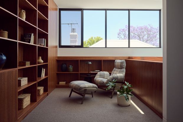 A compact, warmly furnished reading nook or home office with extensive dark timber built-in shelving and low cabinetry, a large horizontal black-framed window, and a mid-century lounge chair with matching ottoman. Neutral carpet, wood panelling, curated shelving, and a potted plant create a quiet, refined interior. A neighbouring roof and trees are visible outside the window.