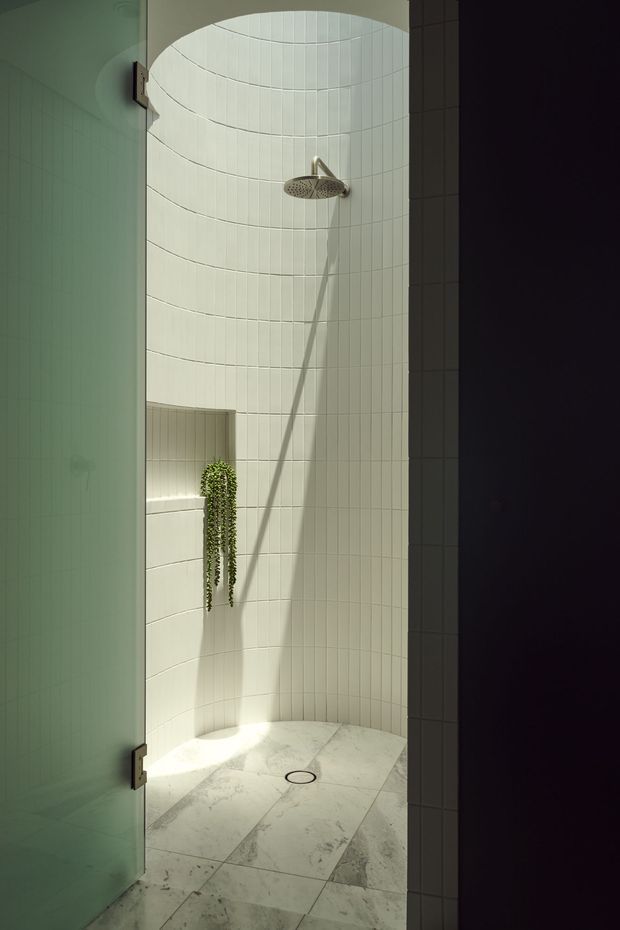 A compact curved walk-in shower enclosed by a frosted glass door. The walls are lined with narrow vertical white tiles following the rounded form, with a rain showerhead, recessed shelf with trailing greenery, and large pale marble-look floor tiles. Soft natural light creates a calm, spa-like atmosphere.