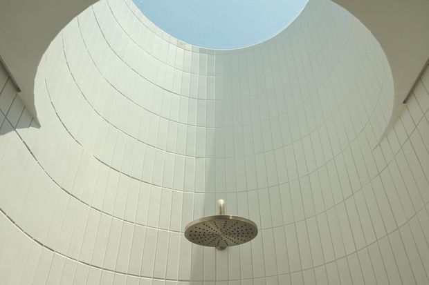 Upward-looking view of a minimalist tiled shower enclosure beneath a large circular skylight or roof opening. The curved walls are clad in pale white vertical rectangular tiles, and a round brushed-metal rain showerhead projects from the centre, creating a bright, clean and sculptural composition.