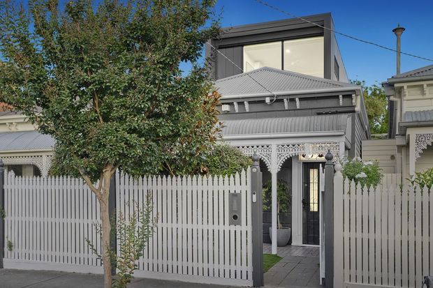 Street-facing view of a compact contemporary extension set behind a traditional ornate Victorian-style façade. The property features a grey corrugated metal roof, decorative lacework verandah detailing, pale picket fencing, a dark-framed glazed upper level, and a paved entry path with established planting.