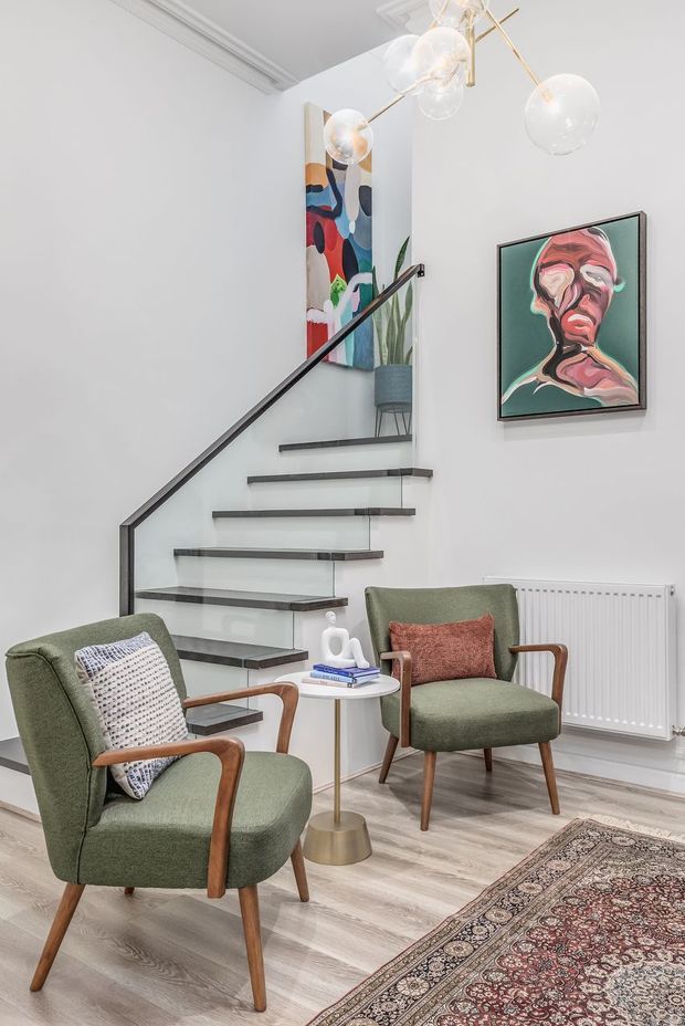 Bright, refined stair hall and sitting area with a floating open-riser staircase, dark slim handrail, pale timber flooring, two green upholstered mid-century armchairs, a small round side table, framed abstract artwork, a radiator, patterned rug, and sculptural pendant lighting. Decorative ceiling mouldings and a high stair void add architectural character.