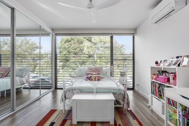 Bright children's bedroom with a white metal bed, colourful striped rug, white storage shelving, mirrored wardrobe doors, and full-height glazed doors opening to a balcony. The room has light timber-look flooring, white walls, a ceiling fan, and an air-conditioning unit.