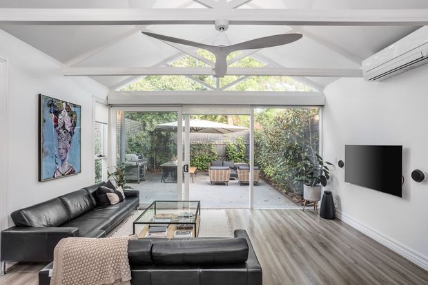 Bright contemporary living room with a black leather sectional sofa, glass coffee table, wall-mounted television and speakers, light timber-look flooring, and white walls. Exposed white roof trusses and a ceiling fan create a high vaulted ceiling, while large sliding glass doors open to a landscaped outdoor patio.