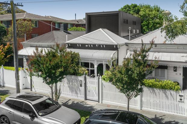 Street-level exterior view of a predominantly white, ornate residential house with a corrugated metal roof, decorative veranda, white picket fencing, and landscaped frontage. A dark contemporary box-like upper extension is visible behind the traditional roofline, with neighbouring homes and parked cars in the foreground.