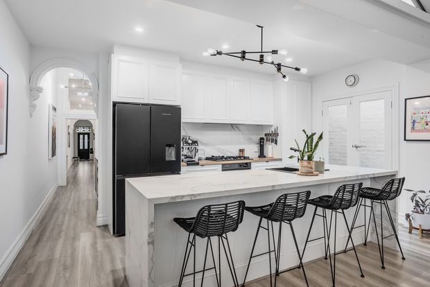 Bright, spacious white kitchen with a large marble-look island and five black woven bar stools. The room features white shaker-style cabinetry, a large matte-black refrigerator, light wood-look flooring, black modern track lighting, and glazed double doors. An arched opening leads to a hallway.