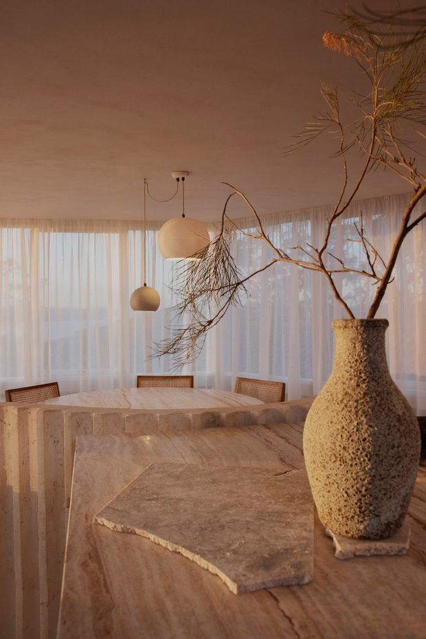 Warm, atmospheric dining room with a large sculptural stone dining table and matching stone slab surfaces. Sheer floor-to-ceiling curtains wrap the room, while woven-back dining chairs and two simple spherical pendant lights create a soft, understated setting. A tall textured ceramic vase with branching foliage forms a prominent foreground feature.