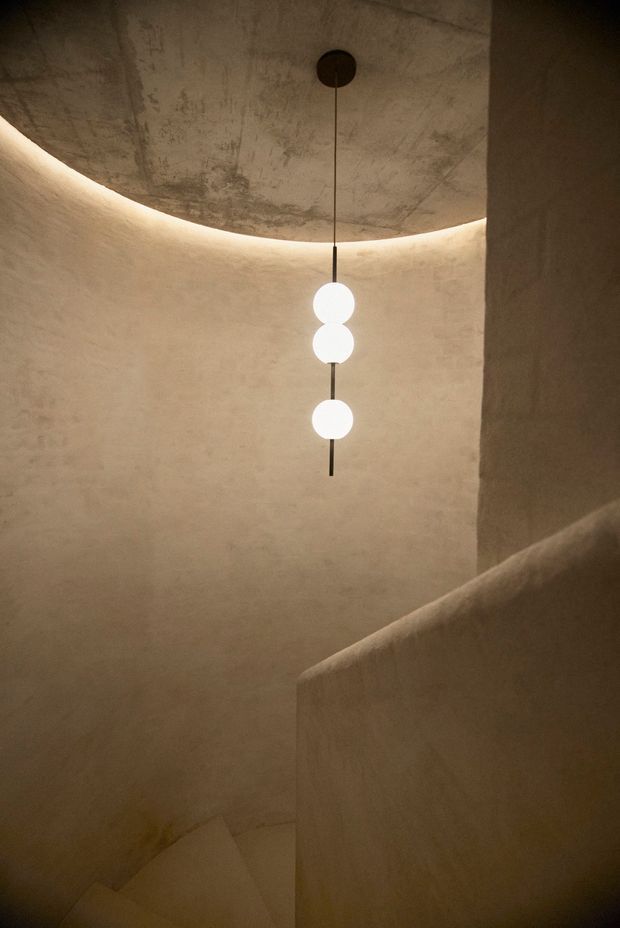 A dimly lit, sculptural stairway with curved plaster walls and a raw concrete ceiling. Warm concealed lighting traces the ceiling edge, while a minimalist pendant with three glowing spherical lights hangs over the stairwell. The stair enclosure has a soft, monolithic, textured finish and a dramatic contemporary atmosphere.
