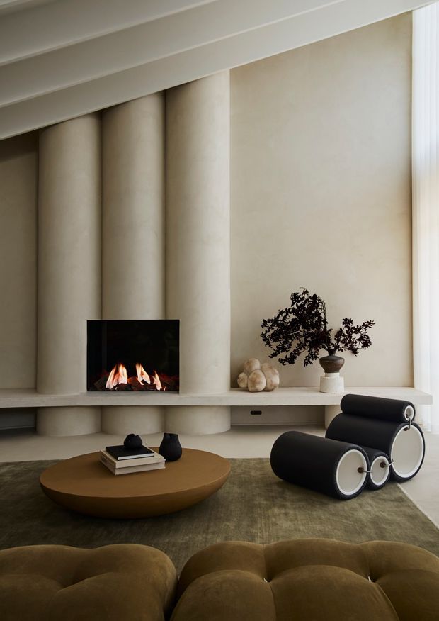 A refined contemporary living room centred on a sculptural fireplace wall with tall rounded vertical plaster columns and a long integrated hearth shelf. The room includes a low rounded timber coffee table, a distinctive black-and-white lounge chair, a deep olive-brown tufted sofa in the foreground, muted textured carpeting, and restrained decorative objects. Soft daylight enters through full-height sheer curtains at the right, creating a warm, minimalist and luxurious atmosphere.