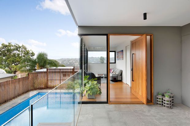 Contemporary covered outdoor terrace beside a swimming pool, opening into a bright interior living space through large glazed doors. The scene features grey tiled flooring, a frameless glass pool balustrade, timber fencing, a timber-framed pivot door, and warm timber flooring inside. A leafy suburban hillside setting is visible beyond the pool.