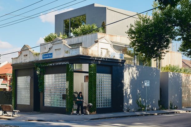 Street-facing view of a contemporary multi-level building incorporating a dark, older-looking ground-floor commercial facade with extensive glass-block glazing, green tiled columns, signage, and a small entry. A modern light-grey upper-level addition has a boxy flat-roof form, large window, rooftop planting, and vertical timber-like cladding. The building sits directly on a leafy urban streetscape with overhead power lines and a footpath.