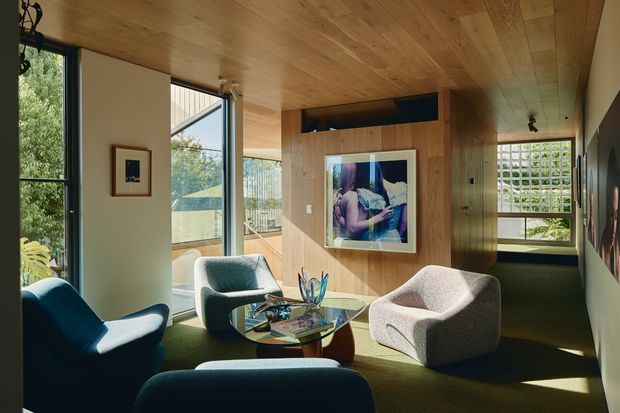 A warm, wood-lined sitting room with expansive glazing, a low built-in window seat, sculptural upholstered lounge chairs, and a glass coffee table. Natural light enters through the windows and a patterned masonry screen, while framed artwork and dark green carpet add contrast.