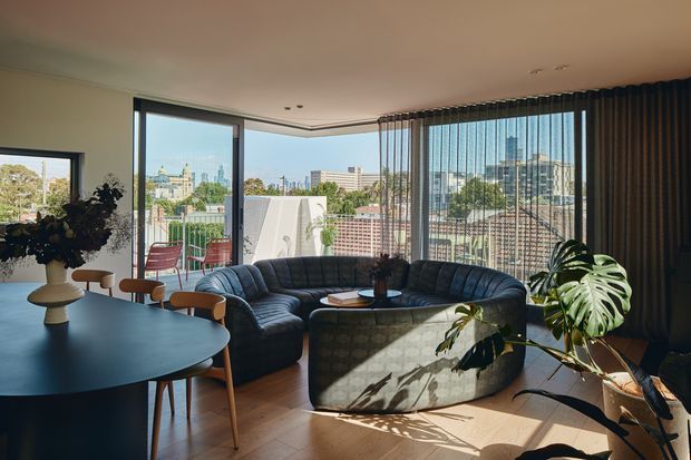 A bright, contemporary open-plan living and dining area with expansive corner glazing and a city outlook. A large curved dark sectional sofa surrounds a matching rounded coffee table, while a dark oval dining table with timber chairs occupies the foreground. Full-height vertical blinds, timber flooring, indoor plants, and a balcony with outdoor seating are visible.