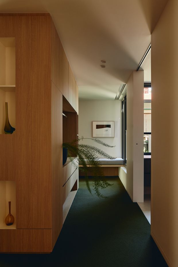 A narrow, warmly lit residential hallway leading to a built-in window seat. Full-height timber veneer cabinetry lines the left wall, incorporating open display niches and concealed storage. A dark green carpet runs through the corridor, with pale walls, a framed artwork, a potted fern, and a glazed opening bringing in daylight at the far end. The space has clean, tailored joinery and a restrained contemporary feel.