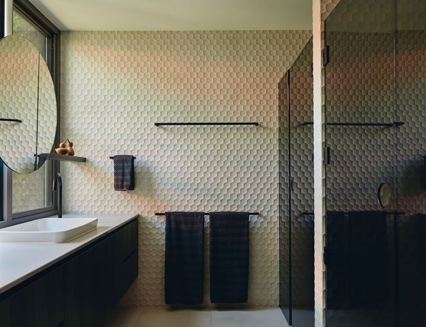 Contemporary bathroom with a floating dark vanity, white integrated basin, round wall mirror, wall-mounted tapware, heated towel rail with dark towels, and a frameless glass shower enclosure. The walls are clad in a distinctive three-dimensional honeycomb-patterned tile, creating a textured feature surface. Natural light enters through the side window, while slim linear wall lighting provides task illumination.