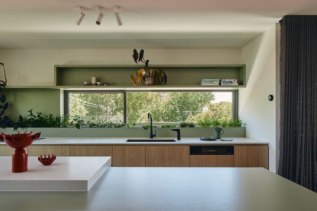 A contemporary kitchen viewed from the main work area, featuring a long wall of light timber cabinetry, a dark benchtop with integrated sink and tapware, a large white island, and a broad horizontal window overlooking greenery. An open recessed display shelf runs above the window, with indoor plants and restrained decorative objects. The room has a pale neutral palette, clean lines, track lighting, and a dark full-height curtain at the right.