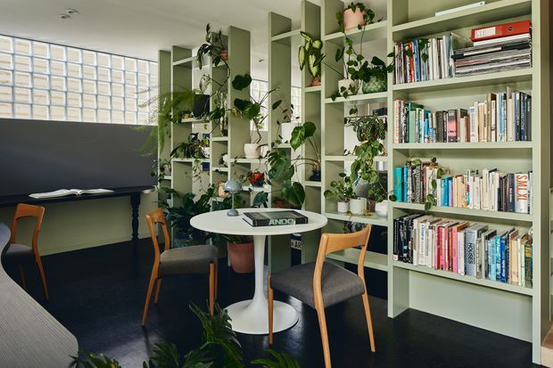 A bright, plant-filled interior combining a compact dining or meeting area with extensive floor-to-ceiling built-in shelving. Pale sage-green open shelving displays books, objects, and indoor plants, while a small round white pedestal table is surrounded by timber-framed upholstered chairs. A dark charcoal wall and dark floor contrast with the light joinery, and a glass-block window provides diffuse daylight.