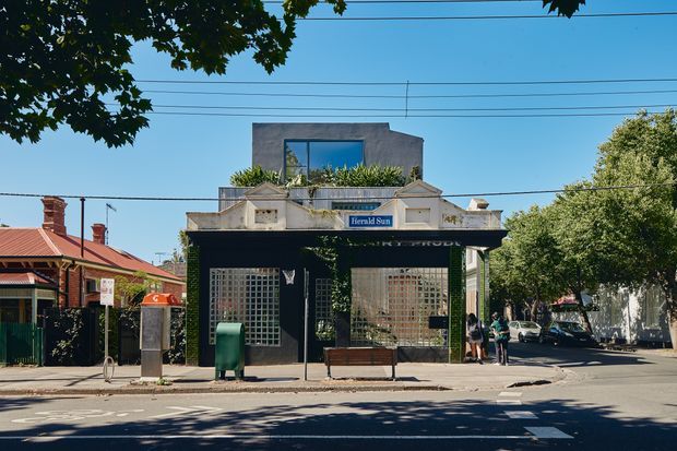 Street-facing view of a small urban commercial building with a dark ground-floor shopfront, glass-block window screens, a sheltered entrance, and a rooftop box-like extension with large glazing. The building sits within an established residential streetscape, with overhead utility wires, mature trees, adjacent heritage-style houses, and clear daylight.