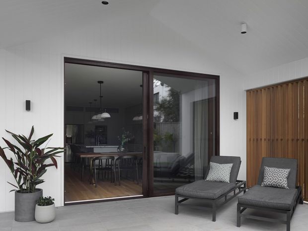 A contemporary covered outdoor terrace or patio with pale tiled flooring, white vertical wall cladding, dark-framed full-height sliding glass doors, and a timber slatted privacy screen. Two dark outdoor lounge chairs with footstools sit in the foreground, while a dining area and kitchen are visible through the glazing. Potted plants and reflected greenery add natural accents.