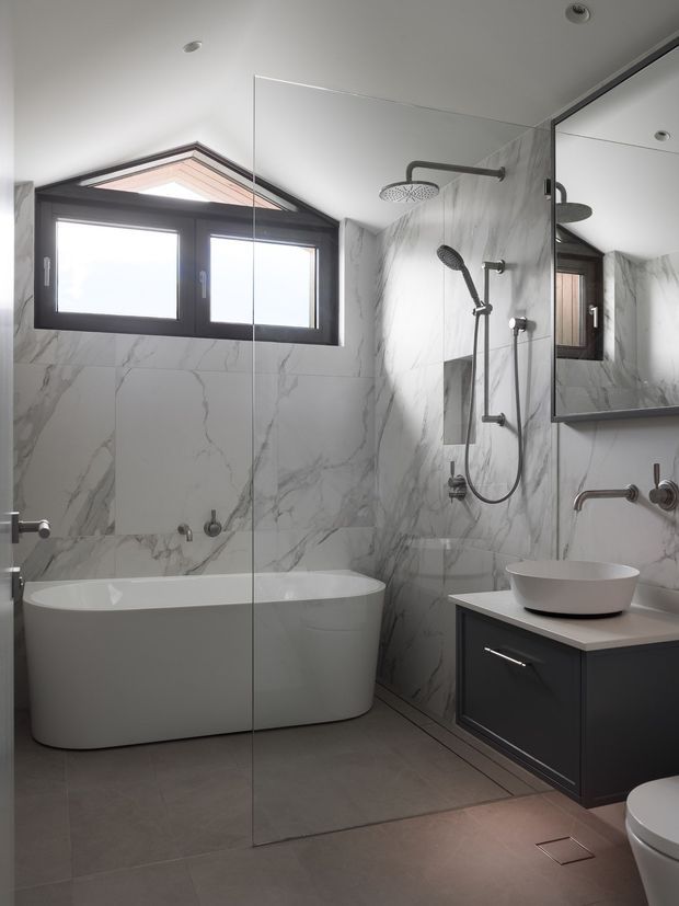 A contemporary bathroom with a freestanding white bathtub, walk-in shower behind a frameless glass screen, wall-mounted shower fittings, vessel basin on dark vanity cabinetry, large mirrored cabinet, marble-look wall tiles, and large grey floor tiles. A dark-framed window provides natural light.