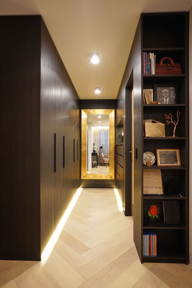 A narrow, warmly lit hallway lined with dark timber built-in cabinetry and open display shelving. Pale herringbone timber flooring contrasts with the dark joinery, while concealed linear lighting illuminates the floor edges. A large framed mirror at the end of the corridor reflects a softly furnished room beyond, adding depth and brightness.