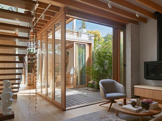 Warm contemporary living room opening onto a small timber-decked courtyard through expansive glazed timber-framed sliding or folding doors. The room features exposed timber ceiling beams, a floating open-riser staircase with timber treads, pale brick walls, timber flooring, a built-in fireplace or media unit, and modern lounge furniture. Strong indoor-outdoor connection with abundant natural light and greenery beyond.
