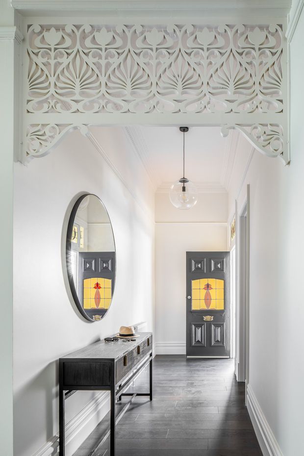 Bright, narrow heritage-style hallway with dark timber-look flooring, white walls and ornate painted decorative fretwork forming a screen above a wide opening. A slim dark console with drawers sits beneath a large oval mirror, while a black door with colourful stained-glass panels anchors the far end. A simple glass pendant hangs from a detailed coffered and corniced ceiling.