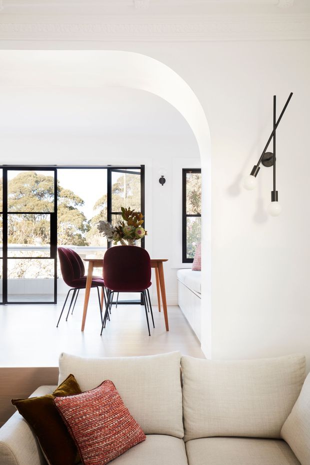 Bright, refined interior view showing a living room in the foreground opening into a dining area. The space features white walls and a large curved archway, black-framed windows, pale timber flooring, a cream upholstered sofa, a mid-century-style timber dining table, burgundy dining chairs, and a sculptural black wall light. Trees are visible through the windows.
