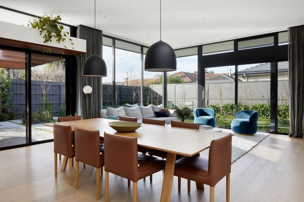 Bright contemporary open-plan dining and living space with a large light timber dining table surrounded by tan upholstered chairs. Floor-to-ceiling black-framed glazing opens to a landscaped courtyard, while oversized black pendant lights, a neutral sofa, blue lounge chairs, dark curtains, and timber flooring create a refined indoor-outdoor setting.