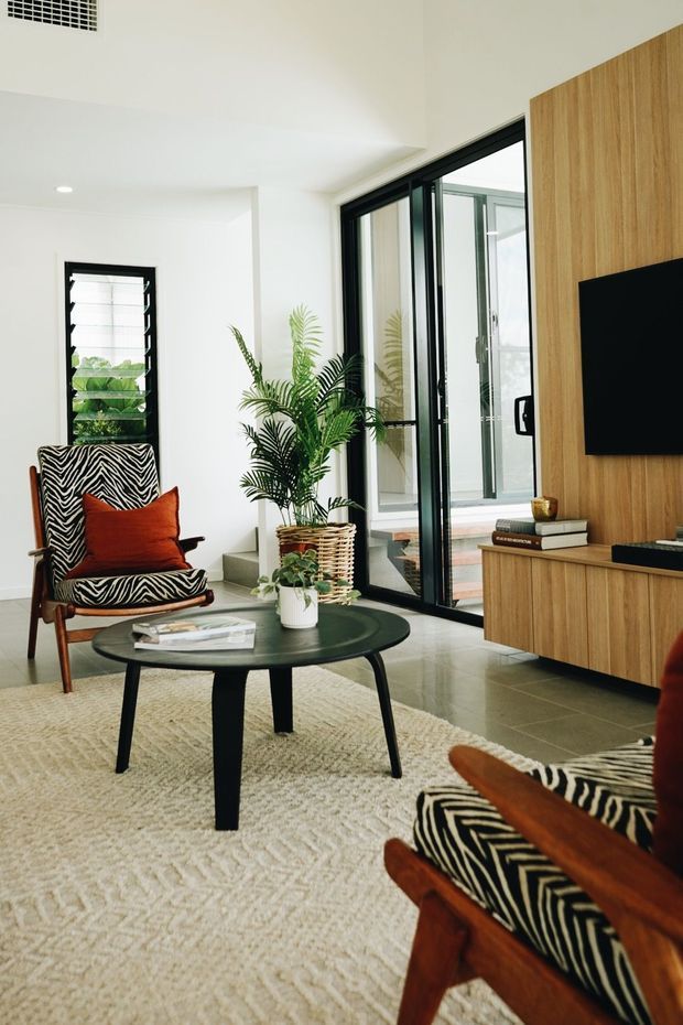 Bright contemporary living room with white walls and ceiling, polished grey concrete-look floor, black-framed glazing and a timber feature wall with matching built-in media console. Mid-century-style timber lounge chairs upholstered in bold zebra print surround a dark round coffee table, with a textured cream rug, terracotta cushion, books and indoor palms. The room opens toward a glazed outdoor area or courtyard.
