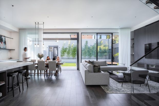 Open-plan contemporary interior combining a kitchen, dining area, and living room. The space has dark timber-look flooring, a white kitchen island with stools, a dining table, a pale sectional sofa, large black-framed glazed doors and windows, and a dark built-in media wall with shelving. Full-height glazing opens views to a small landscaped courtyard, while slim pendant lights and recessed ceiling lights create a bright, refined atmosphere.