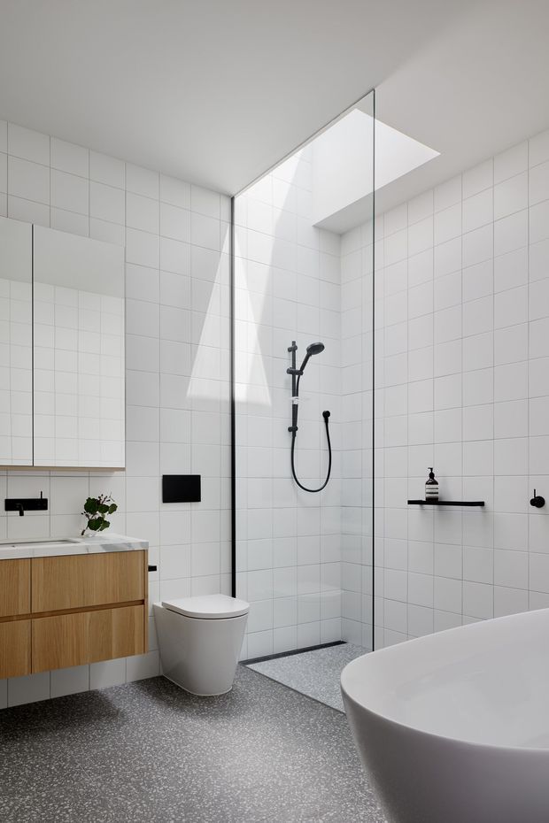 Bright contemporary bathroom with white grid wall tiles, a large frameless glass walk-in shower beneath a skylight, wall-mounted timber vanity, white toilet, and freestanding white bathtub. Dark fixtures and a terrazzo-like grey floor provide contrast.