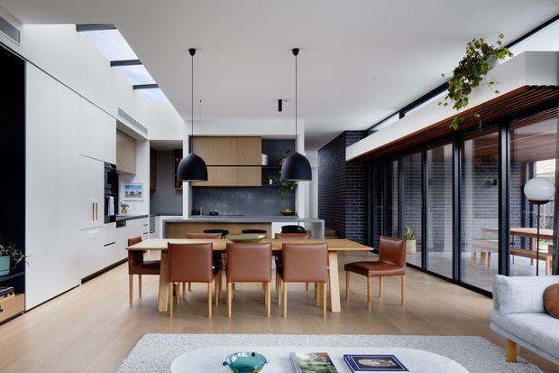 Open-plan contemporary kitchen and dining area with a long timber dining table, leather dining chairs, white integrated cabinetry, a central island, black pendant lights, and extensive glazing with horizontal blinds. A living area is partially visible in the foreground, while a dark textured feature wall and indoor greenery add contrast.