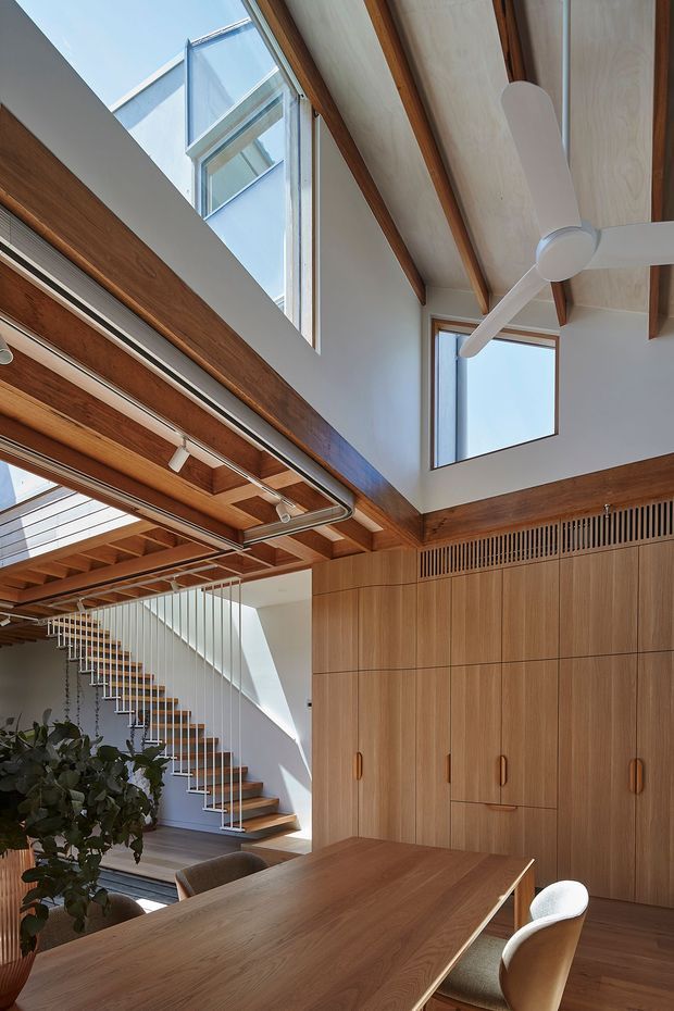A bright, double-height open-plan interior featuring a timber dining table and upholstered chairs in the foreground, a sculptural open-riser staircase with slender vertical balustrades, extensive built-in timber cabinetry, exposed timber beams, clerestory glazing, and a large white ceiling fan. The space has strong natural light and a warm, refined timber palette.