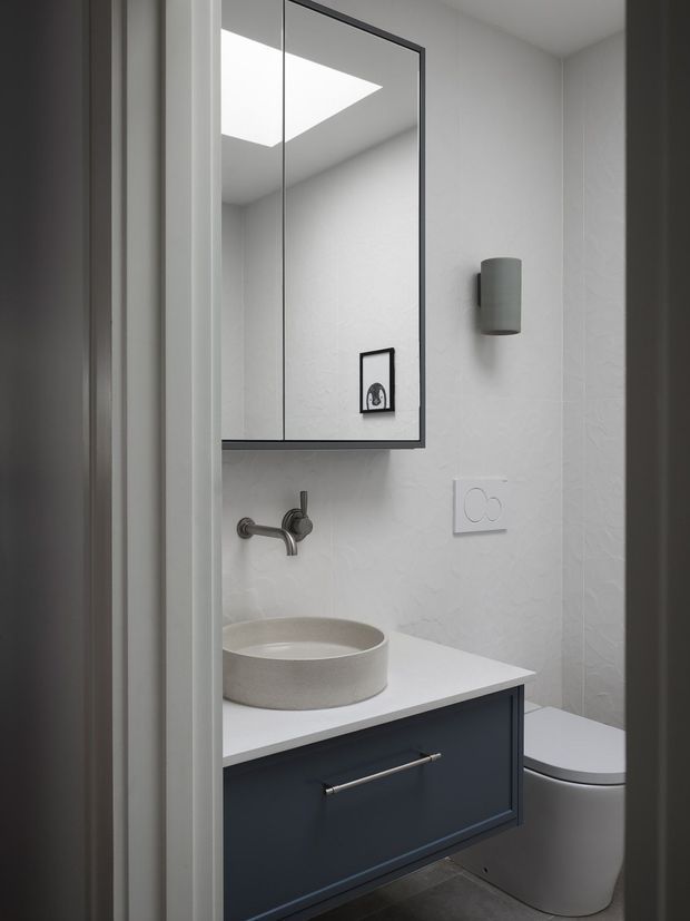 A compact, contemporary bathroom viewed through a doorway, featuring a wall-mounted dark vanity with a white benchtop, round vessel basin, wall-mounted brushed metal tapware, large framed mirror, white textured wall tiles, cylindrical wall light, and a concealed-cistern toilet. A skylight is reflected in the mirror, contributing to the bright, clean atmosphere.