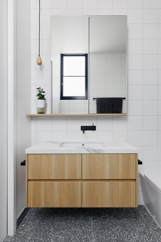 A compact contemporary bathroom with white square tiled walls, a wall-mounted light timber vanity with four drawers, a white stone-look benchtop and integrated basin, and matte black tapware. A large mirrored cabinet spans the wall above the vanity, with a small black-framed window, floating timber shelf, dark towel, pendant light, and potted plant. The floor is finished in dark terrazzo-style material, and a bathtub is partially visible at right.