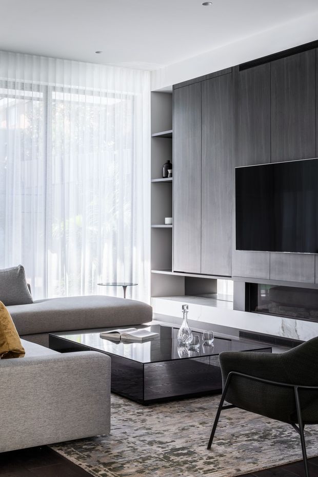 A refined contemporary living room with a large grey sectional sofa, black lounge chair, glossy dark coffee table, and patterned area rug. Floor-to-ceiling sheer curtains provide abundant natural light, while a full-height dark timber entertainment wall incorporates open shelving, a television, and a linear fireplace with a pale stone surround. The room has a clean, sophisticated, monochromatic palette with warm timber accents.