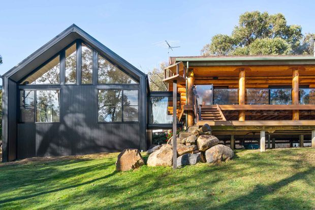 Exterior view of a rustic timber cabin-style dwelling beside a modern dark corrugated-metal gabled structure. The timber building has exposed round log posts, a covered elevated veranda, stairs, and large glazed openings, while the adjacent structure features a steep gable roof and extensive dark metal cladding and glazing. The buildings sit in a grassy landscaped setting with large rocks and mature trees.