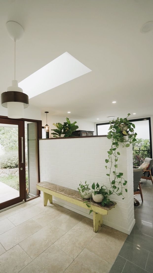 Bright residential entryway with a white painted brick half-height partition wall, dark timber-framed glazed doors and windows, pale stone or porcelain tiled flooring, and a rustic timber bench decorated with potted trailing plants. A large pendant light and recessed ceiling lights illuminate the space, with a furnished living area visible beyond the partition.