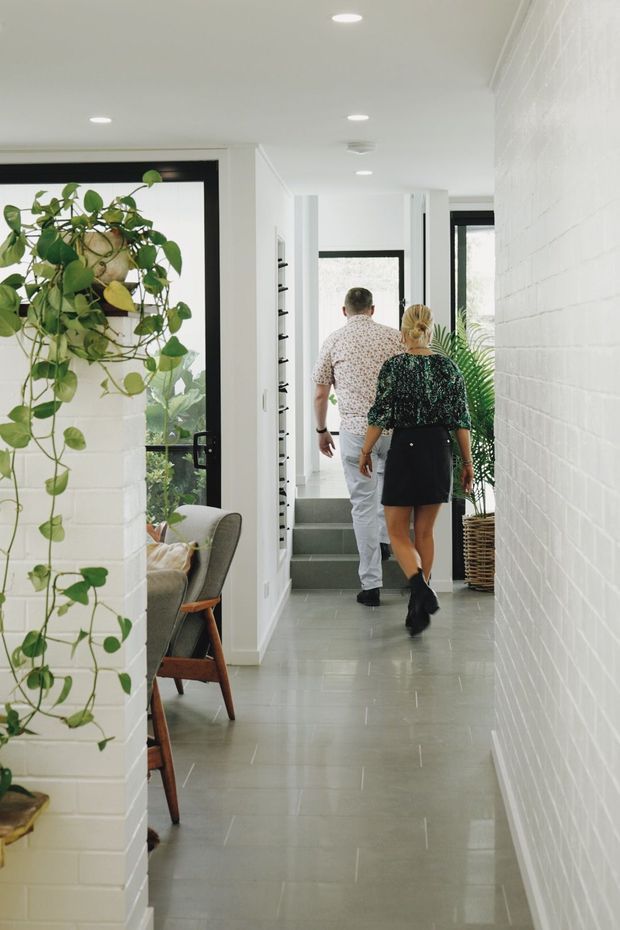A bright, narrow residential hallway with a long grey tiled floor, white-painted brick walls, recessed ceiling lights, and black-framed glazed doors and windows. A leafy indoor plant and partial mid-century-style seating are visible on the left, while the corridor leads to a short flight of steps and additional glazing at the rear. Two people are walking through the space.