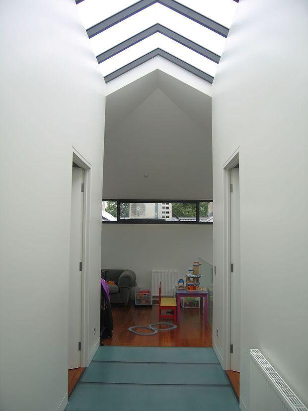 A bright, minimalist interior hallway with tall white walls and a dramatic pitched ceiling incorporating multiple linear skylights. The corridor has white doorways on both sides, a dark teal or blue-green floor, and opens into a children's play or living area with timber flooring, a sofa, toys, and low tables. High clerestory-style glazing at the far end provides views of nearby urban buildings.