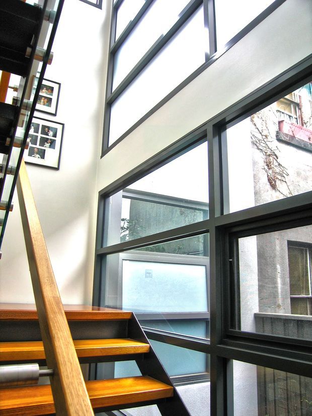 Interior view of a modern stairway with open timber treads, a timber handrail, dark structural supports, and extensive floor-to-ceiling glazing. The double-height stairwell is bright and airy, with framed photographs on the wall and urban buildings visible outside.