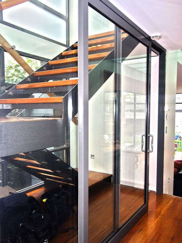 Interior view of a modern stairway with open-riser timber treads supported by dark structural elements and enclosed beside a full-height black-framed glass partition and door. The space has warm timber flooring, extensive glazing, exposed structural framing, and a bright contemporary feel. A small portion of an adjoining room is visible through the doorway.