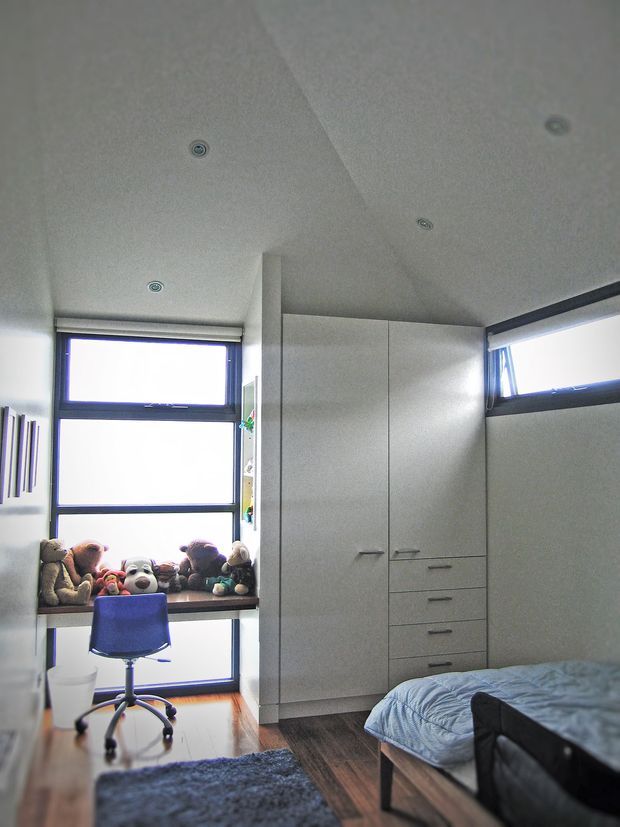 Compact contemporary bedroom with a built-in wardrobe, chest of drawers, desk and blue office chair. Large black-framed windows provide abundant daylight, while a sloped white ceiling with recessed downlights and dark timber flooring define the room. A bed, rug and soft toys are visible.