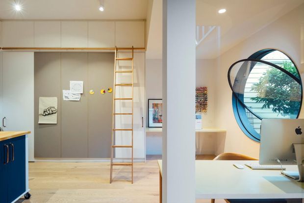 Bright contemporary interior showing a compact built-in study and storage area with a tall timber ladder, pale timber flooring, full-height grey cabinetry, and overhead white cupboards. A blue built-in cabinet and desk occupy the foreground, while a large circular window and iMac define the adjoining workspace. The room has warm recessed lighting and playful contemporary detailing.