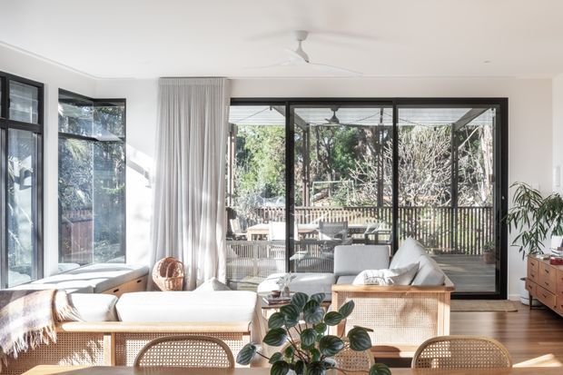 Bright contemporary living room with white walls, warm timber flooring, a rattan-fronted sofa or cabinet, woven furniture, and extensive black-framed glazing opening to a leafy outdoor deck. Sheer curtains, indoor plants, and a ceiling fan create a relaxed indoor-outdoor atmosphere.
