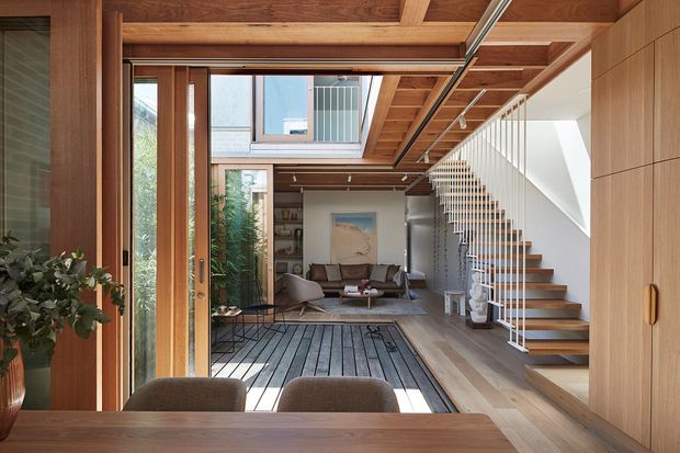 A refined open-plan residential interior viewed from a timber dining area toward a sunlit living room. Warm timber cabinetry, wall panelling, exposed ceiling beams and a slatted overhead structure frame large glazed openings. A floating timber staircase with slender white balustrade rises beside the living area, while a dark linear floor treatment leads toward the sofa and artwork. The space combines strong natural light with warm wood and restrained contemporary furnishings.