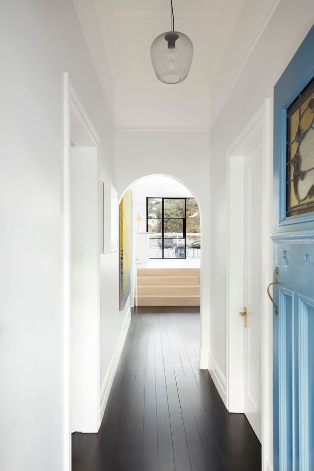 Bright, narrow residential hallway with white walls, decorative ceiling mouldings, white panelled doorways, and dark timber floorboards. A blue decorative glazed front door with stained-glass detailing occupies the right foreground. The corridor ends in an arched opening leading to a light-filled room with a large black-framed window and pale timber built-in cabinetry. A textured glass pendant hangs centrally from the ceiling.