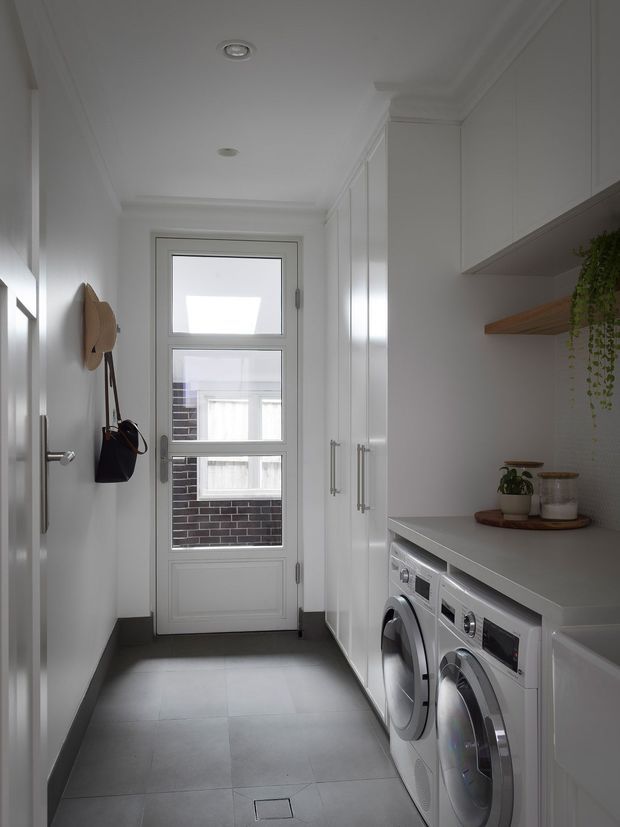 A narrow, bright laundry room with a streamlined built-in layout. White full-height cabinetry lines the right wall above and beside a pale benchtop, with front-loading washing and drying machines installed underneath. A small timber shelf displays potted plants and storage containers. The room has large dark grey floor tiles, white walls and ceiling with recessed lighting, and a glazed white-framed exterior door at the far end. Hooks on the left wall hold a hat and bags.