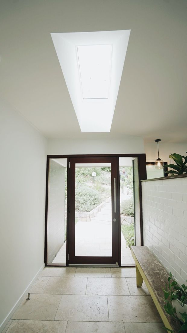 Bright residential entryway or hallway with a large white skylight, pale stone tile flooring, dark timber-framed glazed double doors opening to a landscaped stairway, and a white painted brick feature wall with a rustic bench and pendant light. The space has a clean, airy, contemporary feel with strong indoor-outdoor connection.